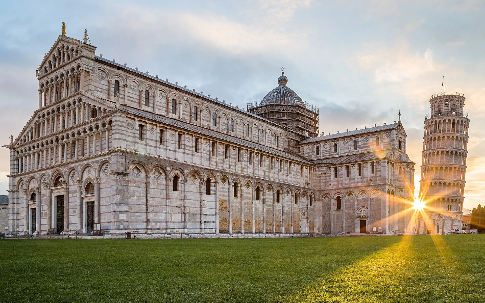The Leaning Tower of Pisa and the marble cathedral on the Piazza dei Miracoli at sunset — a sunburst between them.